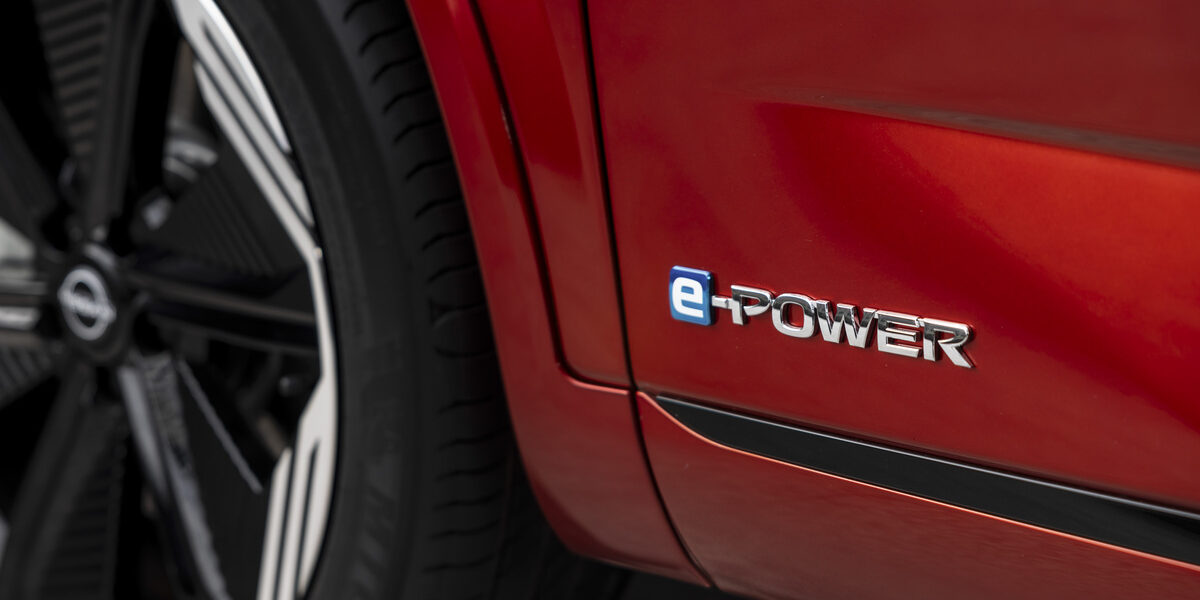 Vue rapprochée d'un véhicule Nissan rouge équipé de la technologie e-POWER, mettant en avant le logo e-POWER en chrome sur le flanc, avec une roue au design moderne visible à l'arrière-plan.