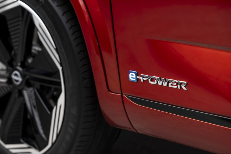 Nissan QASHQAI – e-Power Nissan Troyes Vue rapprochée d'un véhicule Nissan rouge équipé de la technologie e-POWER, mettant en avant le logo e-POWER en chrome sur le flanc, avec une roue au design moderne visible à l'arrière-plan.