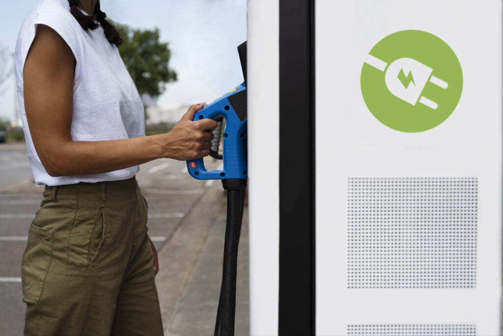 Une personne se tient debout sur un parking et tient une prise de recharge bleue qu’elle est en train de brancher dans une borne de recharge pour véhicule électrique. Elle porte un haut sans manches blanc et un pantalon kaki. Sur la borne blanche, un symbole vert représente une prise électrique avec un éclair, indiquant qu’il s’agit d’une borne de recharge électrique. La scène se déroule en extérieur, par temps clair, avec quelques arbres visibles en arrière-plan.