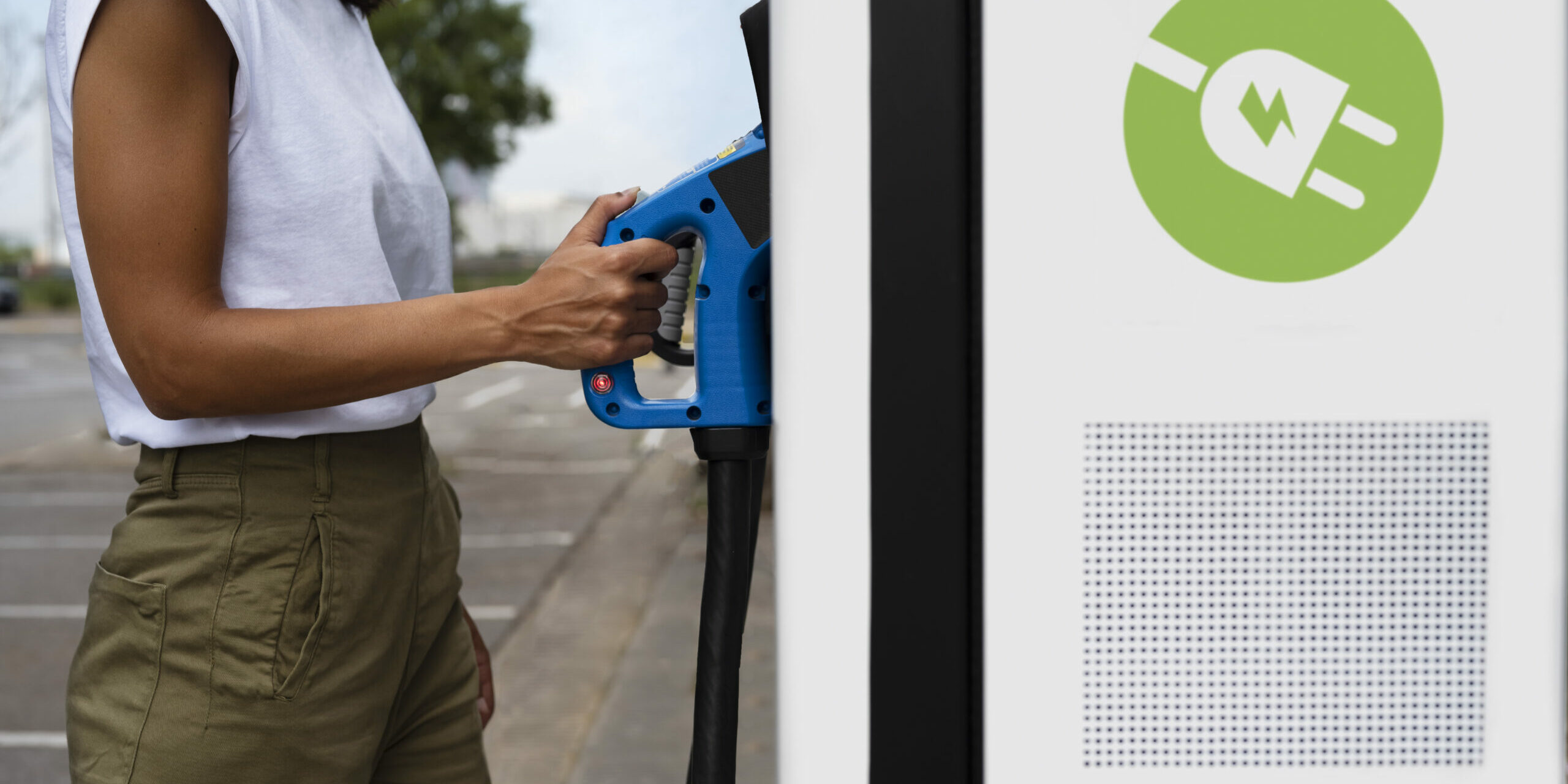 bornes-recharge-troyes Une personne se tient debout sur un parking et tient une prise de recharge bleue qu’elle est en train de brancher dans une borne de recharge pour véhicule électrique. Elle porte un haut sans manches blanc et un pantalon kaki. Sur la borne blanche, un symbole vert représente une prise électrique avec un éclair, indiquant qu’il s’agit d’une borne de recharge électrique. La scène se déroule en extérieur, par temps clair, avec quelques arbres visibles en arrière-plan.