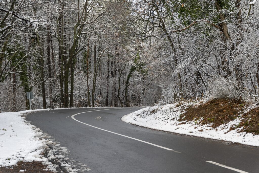 Route sinueuse bordée de forêt enneigée : la chaussée sombre traverse un paysage d’arbres recouverts d’une fine couche de neige, créant un contraste marqué entre l’asphalte humide et les branches blanches. L’atmosphère est calme, hivernale, avec une lumière douce filtrant à travers les troncs.