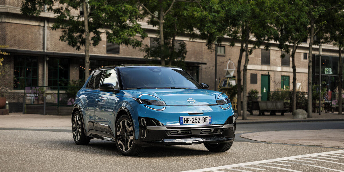 Voiture électrique Nissan MICRA bleue garée sur une place de stationnement en ville, avec des bâtiments en brique et des arbres en arrière-plan. Le véhicule arbore un design moderne et compact
