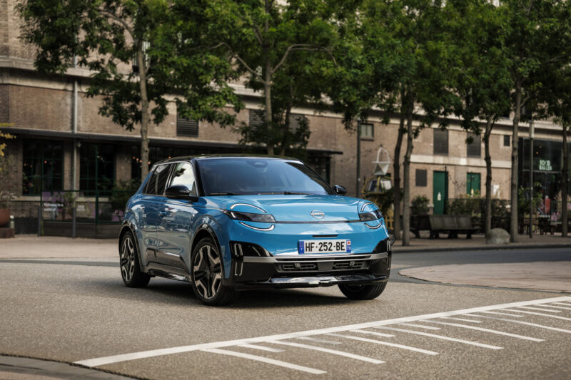 Voiture électrique Nissan MICRA bleue garée sur une place de stationnement en ville, avec des bâtiments en brique et des arbres en arrière-plan. Le véhicule arbore un design moderne et compact